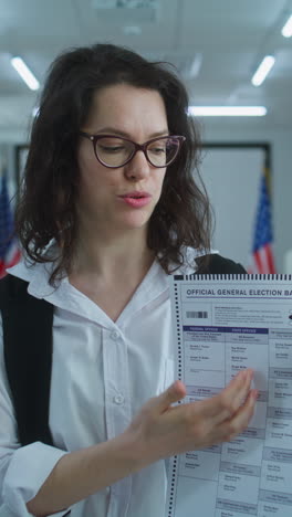 American-woman-speaks-on-camera,-shows-paper-bulletin,-calls-for-voting.-National-Elections-Day-in-the-United-States.-Voting-booths-at-polling-station.-Concept-of-civic-duty-and-patriotism.-Portrait.